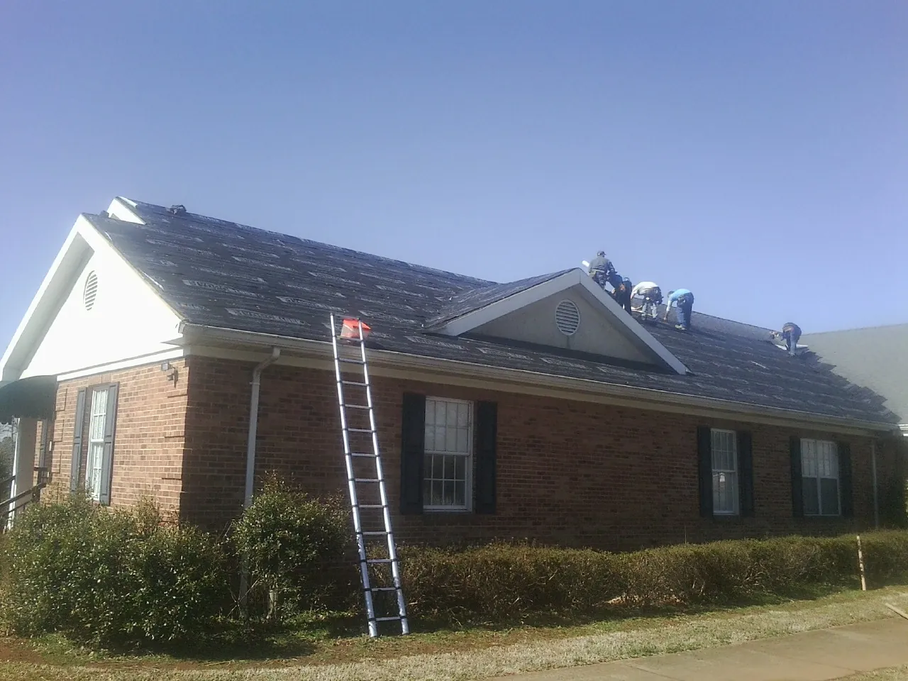 Crew tearing shingles off a brick ranch roof with a ladder set — Metal Roof Repair work by our crew in Mc Coy