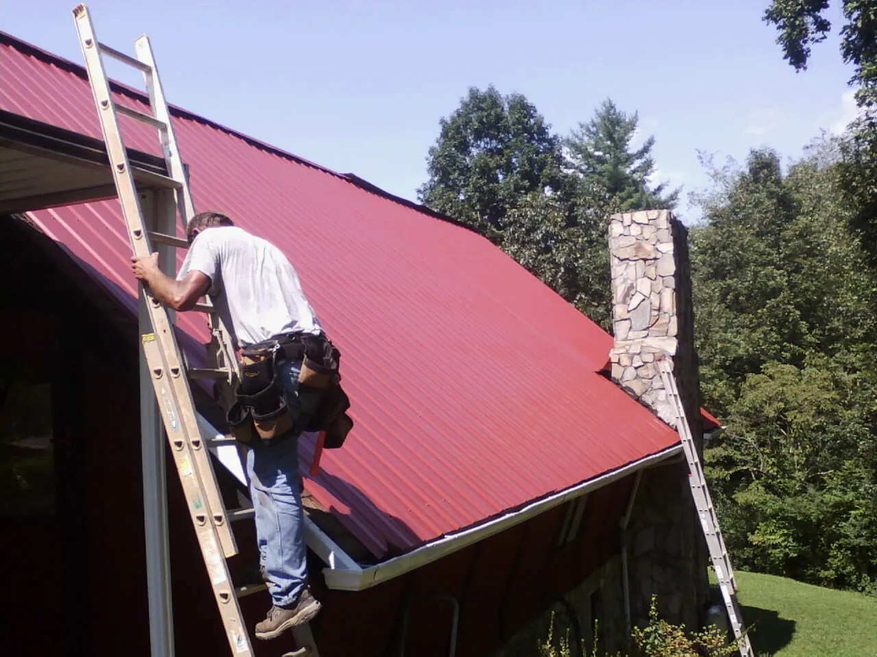 Roofer on a ladder beside a red metal roof and stone chimney — Soffit & Fascia Repair by Roofing Brothers in Mc Coy